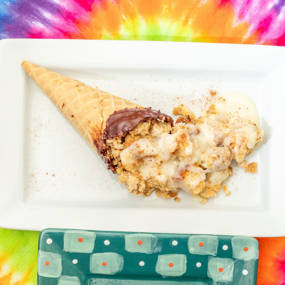 Waffle cone dessert filled with chocolate and crumb topping, drizzled with cream and cinnamon on a white plate.