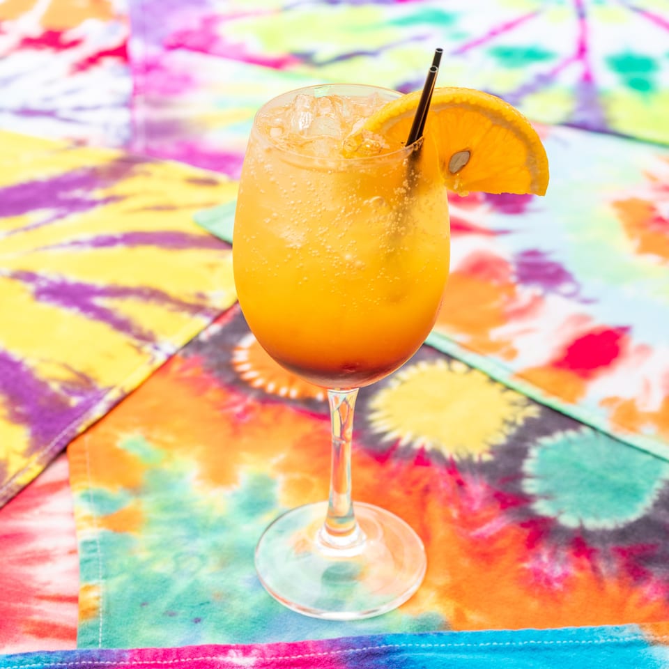 Orange fizzy cocktail with ice, straw, and orange garnish on a vividly colorful painted table.