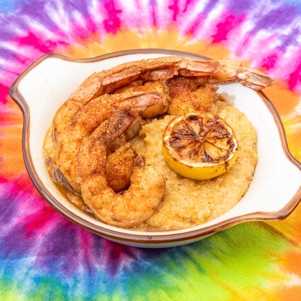 Seasoned shrimp and grilled lemon served over creamy grits in a dish on vibrant tie-dye fabric.