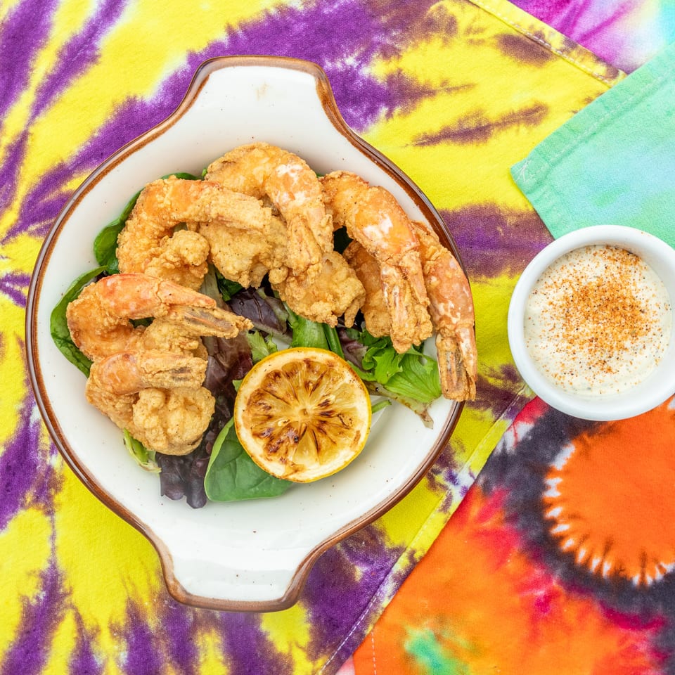 Plate of fried shrimp and seafood over mixed greens with grilled lemon and seasoned dipping sauce on colorful tie-dye fabric.