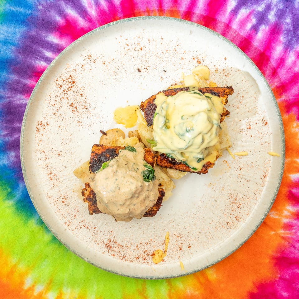 Plated entrée with grilled vegetables, creamy herb sauces, and crispy potatoes on a white plate over colorful tie-dye fabric.