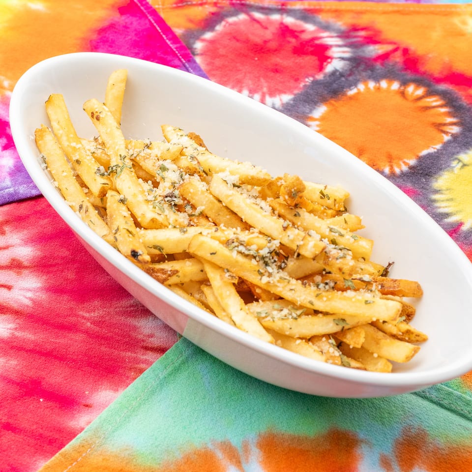 Seasoned fries topped with grated cheese and herbs served in a white dish on vibrant tie-dye fabric.
