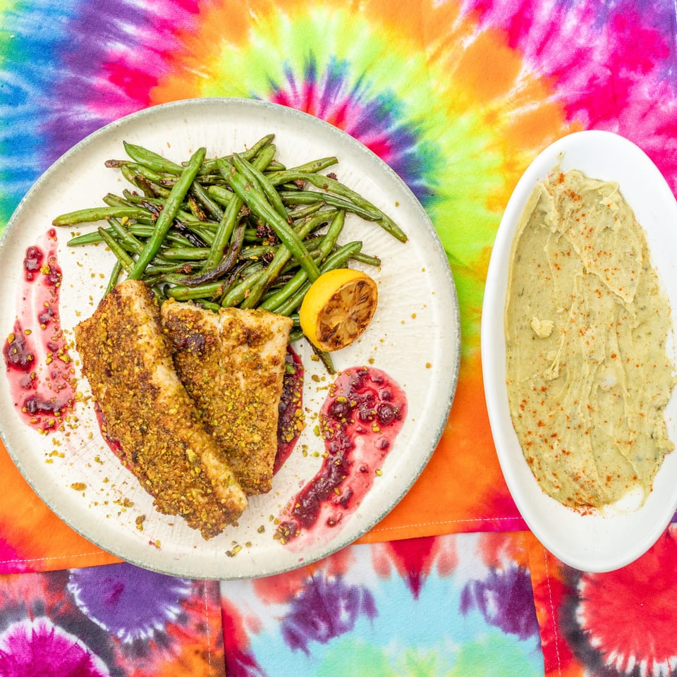 Pistachio-crusted chicken with green beans, grilled lemon, berry sauce, and hummus served on vibrant tie-dye textiles.