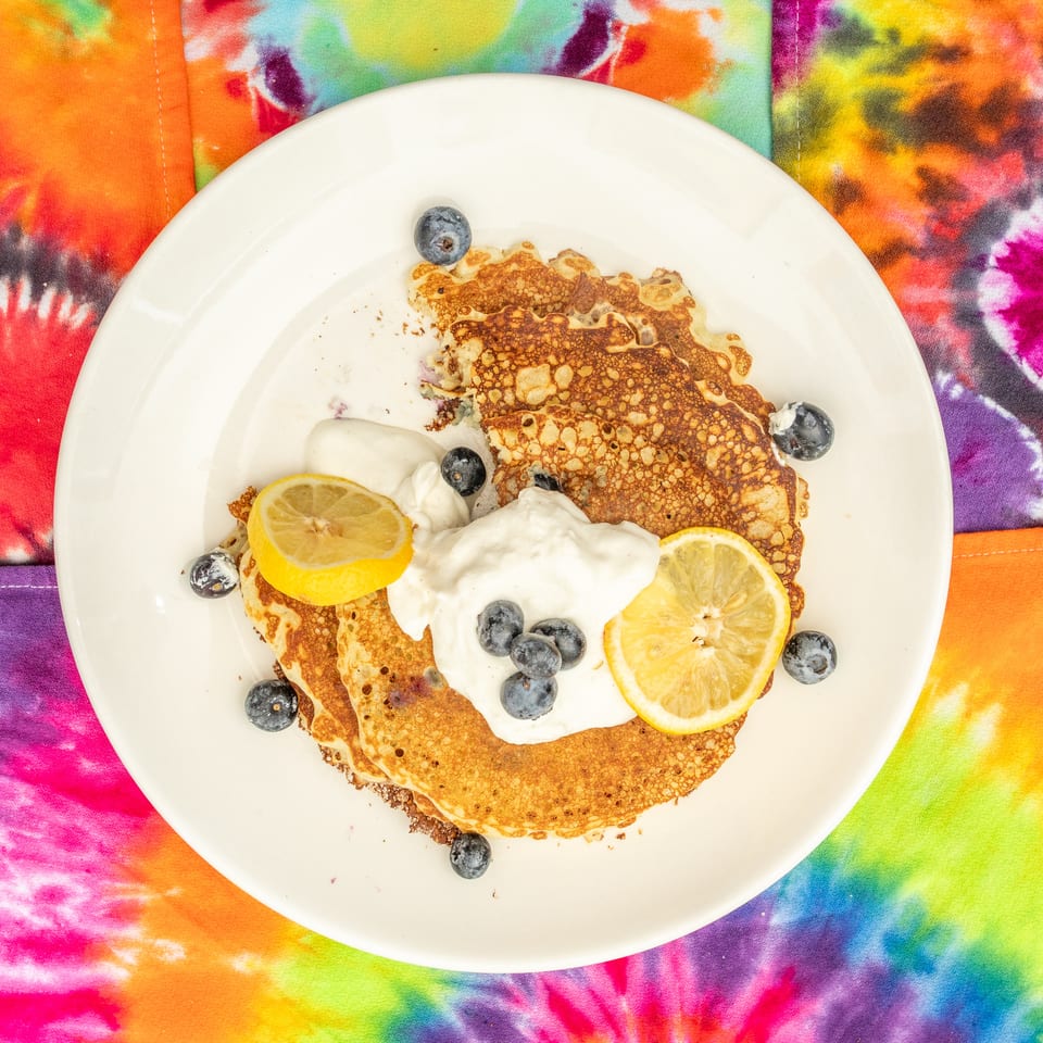 Stack of golden crepes topped with whipped cream, blueberries, and lemon slices on a white plate over vibrant tie-dye fabric.