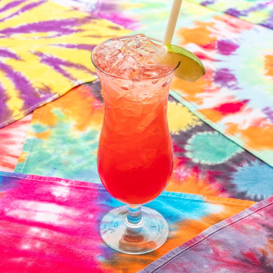 Red iced cocktail with a lime wedge and straw served in a glass over a vibrant tie-dye tablecloth.