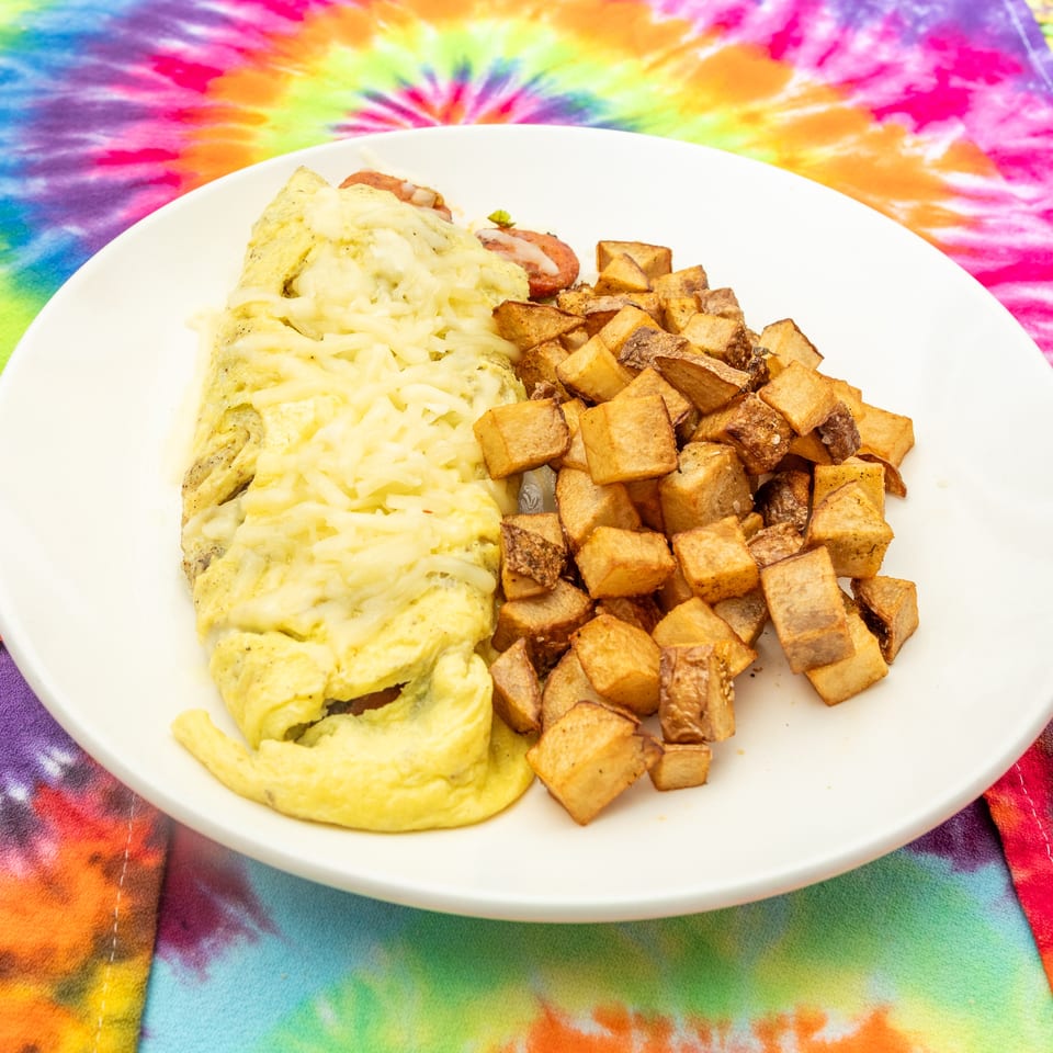 Cheesy herb omelet with roasted diced potatoes on a white plate atop a colorful tie-dye tablecloth.