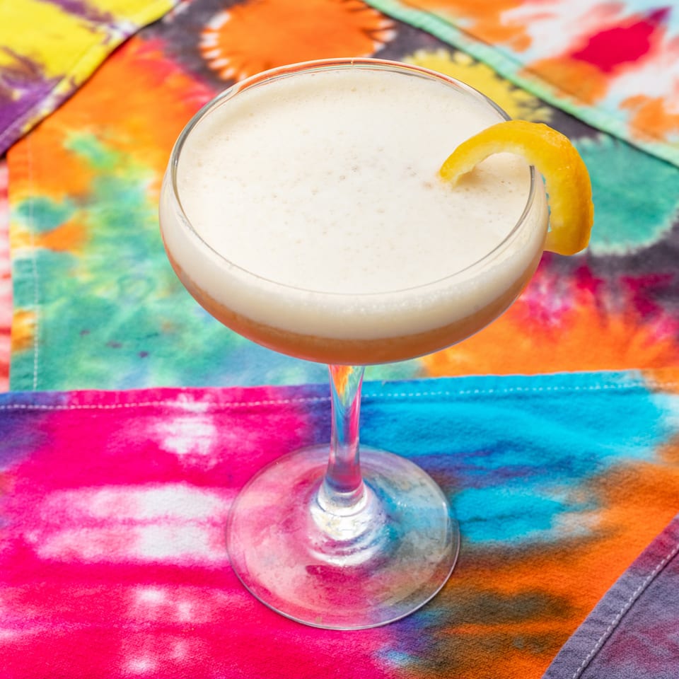 Creamy cocktail in a coupe glass garnished with an orange slice on a vibrant multicolored tablecloth.