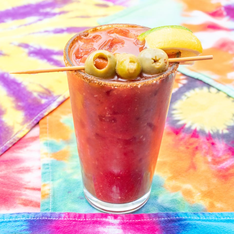 Red cocktail with a chili-salt rim, lime wedge, and skewer of green olives on a colorful tie-dye tablecloth.