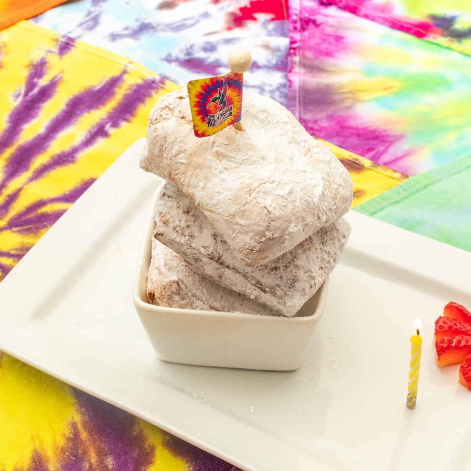 Stack of powdered square pastries in a bowl, topped with a hummingbird flag, alongside strawberries and a lit candle on a colorful tablecloth.