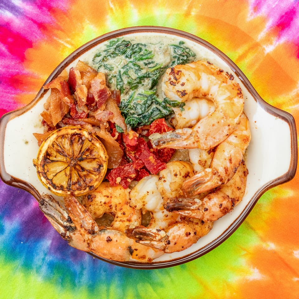 Grilled shrimp platter with creamy spinach, crispy bacon, sun-dried tomatoes, and charred lemon on a colorful tie-dye cloth.