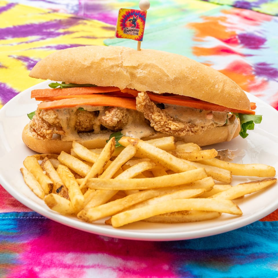Crispy fried chicken sandwich with carrots, greens, and sauce, served with fries on a white plate over a colorful tie-dye tablecloth.