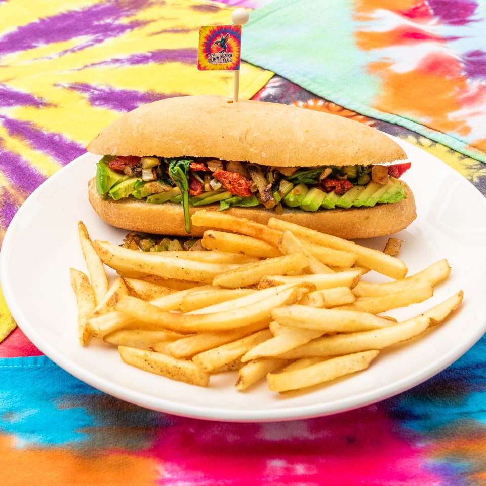 Avocado and vegetable sandwich with fries on a white plate, set against a colorful tie-dye tablecloth.