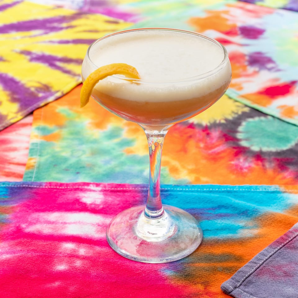 Foamy amber cocktail in a coupe glass garnished with an orange peel, set against a vibrant multicolored tablecloth.