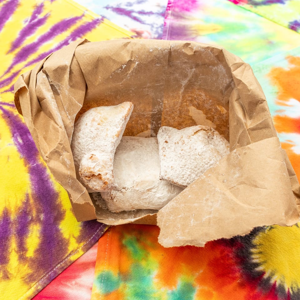 Powdered pastries sit in an open brown paper bag on a vibrant tie-dye cloth.