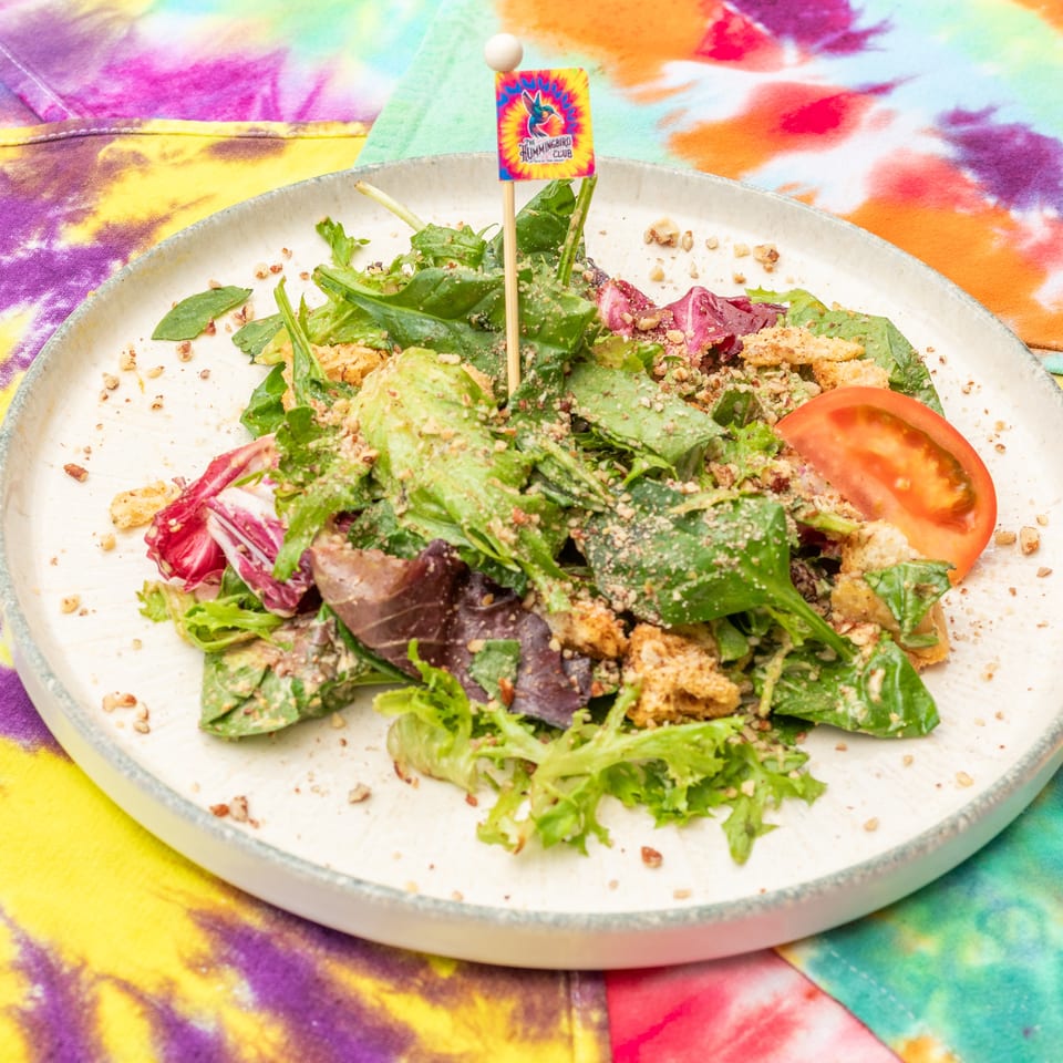 Colorful mixed green salad with tomato, radicchio, crunchy toppings, and a Hummingbird Club flag on a plate.