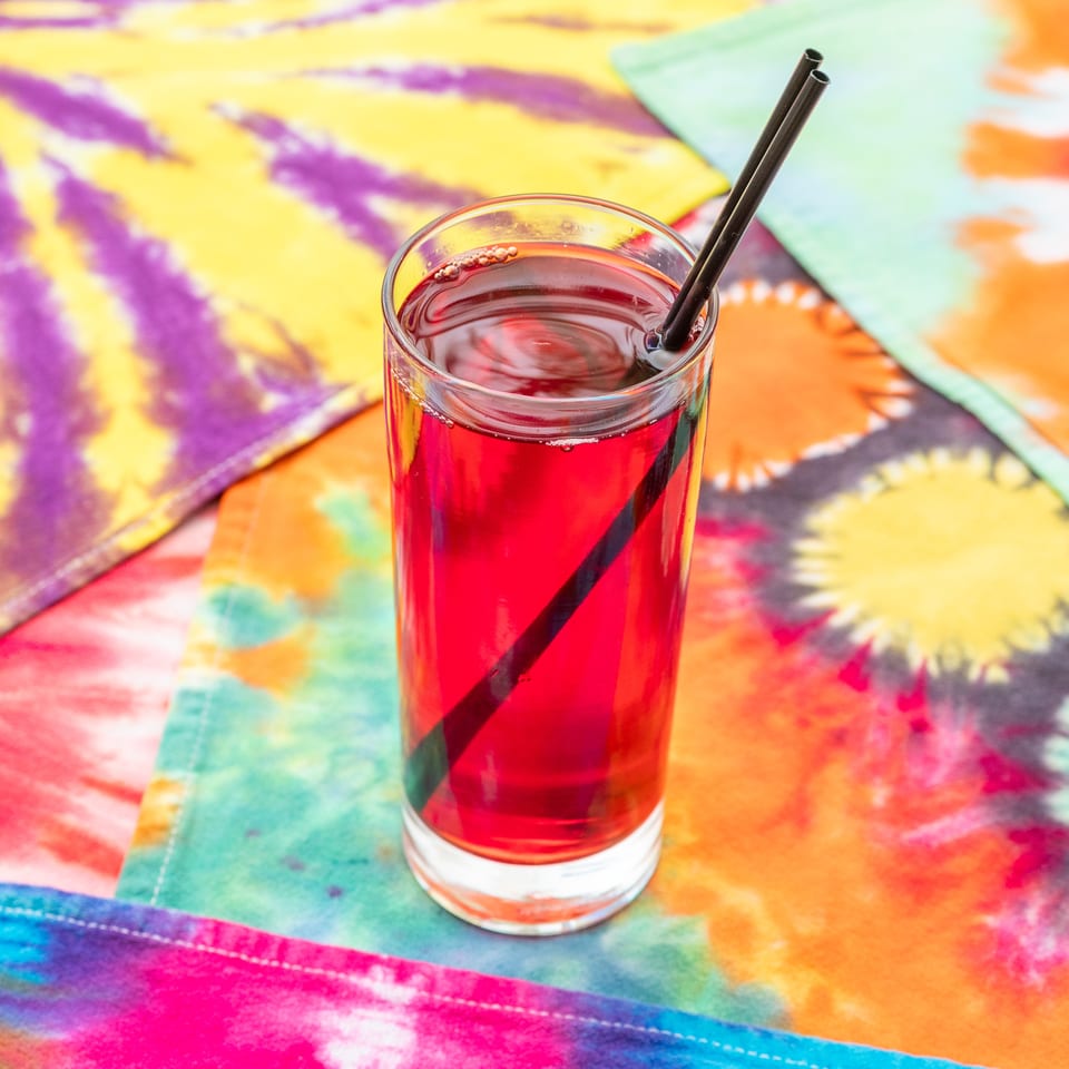 Tall glass of bright red drink with a black straw on a colorful tie-dye tablecloth.