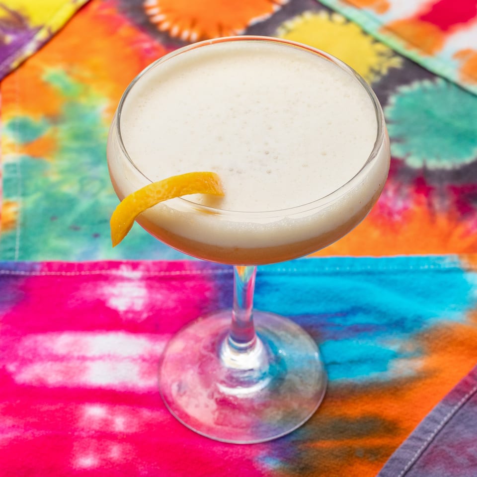 Creamy cocktail in a stemmed glass, garnished with an orange twist, on a vibrant multicolored tablecloth.