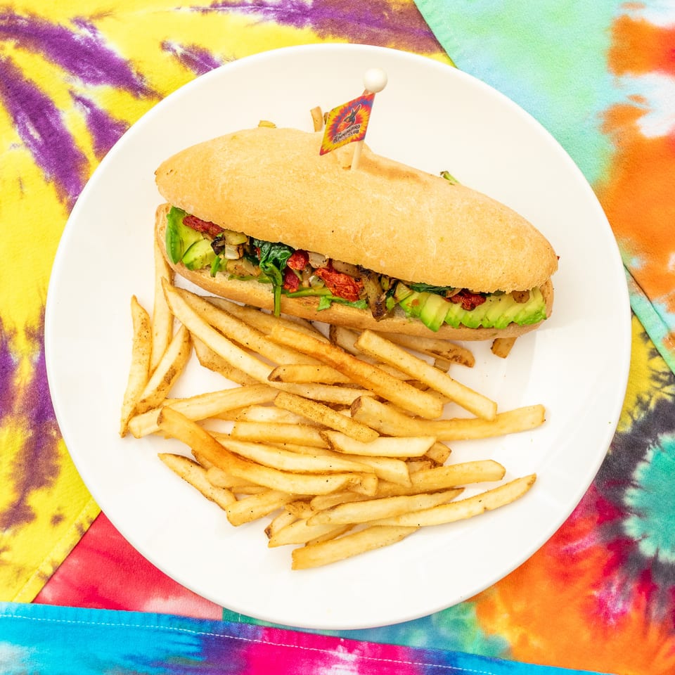 Sub sandwich filled with avocado, vegetables, and sun-dried tomatoes, served with fries on a white plate over colorful tie-dye fabric.