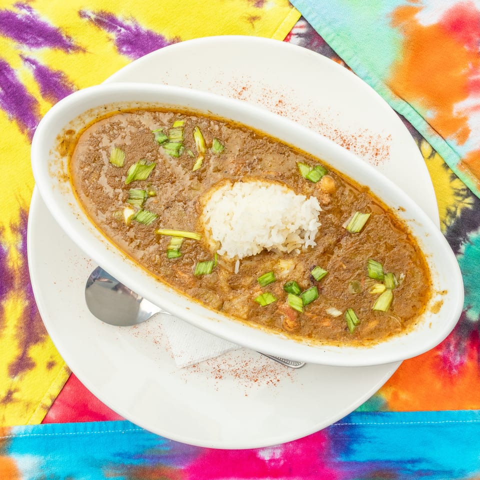 Creamy brown stew topped with white rice and chopped green onions, served in a white dish over colorful tie-dye fabric.