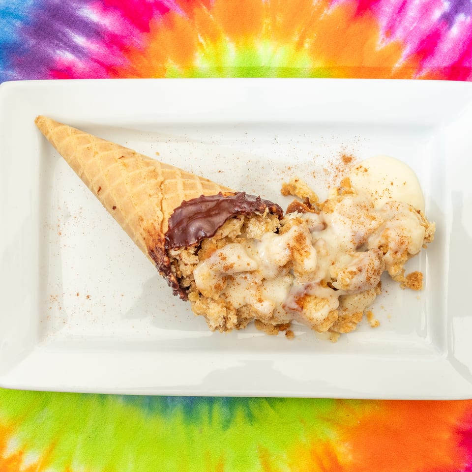 Waffle cone filled with chocolate and vanilla ice cream, topped with crumbled pastry and cinnamon, on a white plate over colorful tie-dye fabric.