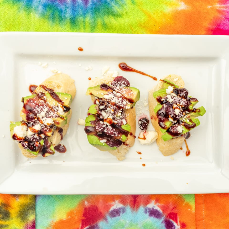 Three avocado-topped crostini drizzled with balsamic glaze and sprinkled with crumbled cheese on a white platter.