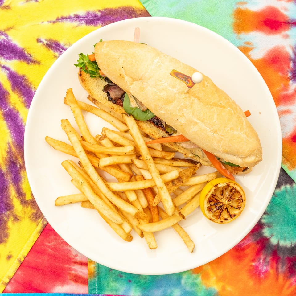 Grilled sandwich with lettuce and vegetables, French fries, and charred lemon on a white plate atop a colorful tie-dye tablecloth.