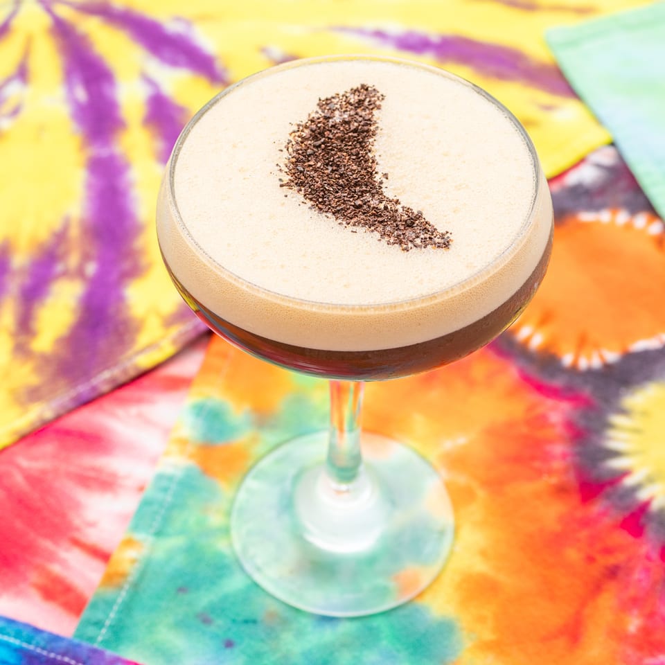 Foamy dark cocktail in a stemmed glass, topped with coffee grounds, against a vibrant multicolored tie-dye backdrop.