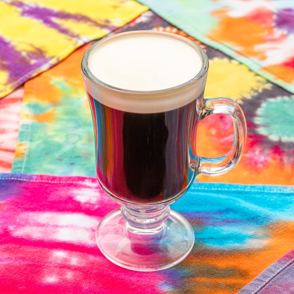 Glass mug of dark coffee topped with creamy foam on a vibrant multicolored tablecloth.