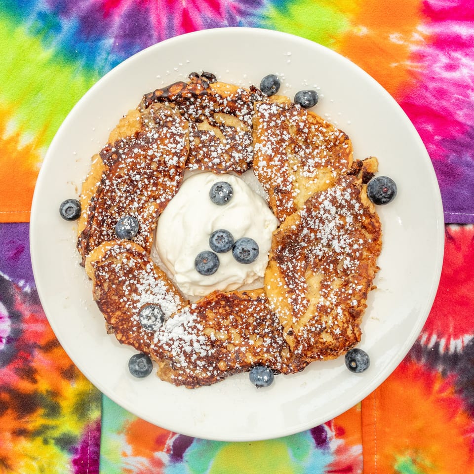 French toast topped with powdered sugar, blueberries, and whipped cream on a white plate over a colorful tie-dye cloth.