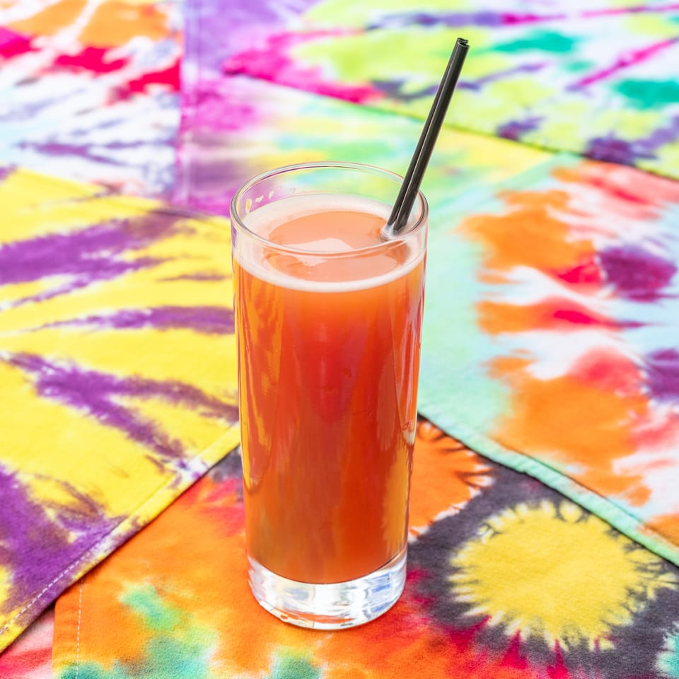 Tall glass of orange-red drink with a black straw on a vibrant multicolored tie-dye tablecloth.