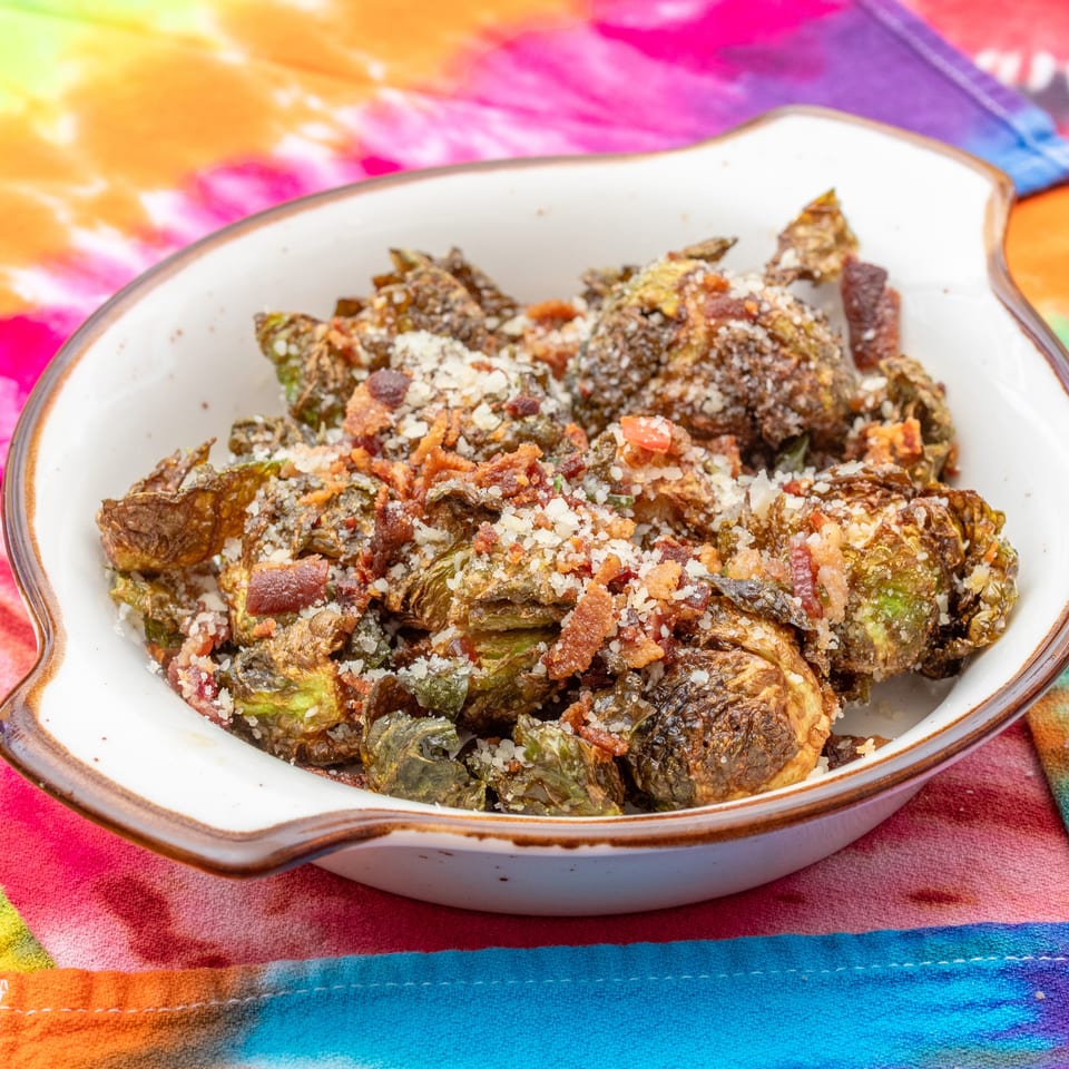 Roasted Brussels sprouts topped with crispy bacon and grated cheese in a white serving dish on a colorful cloth.